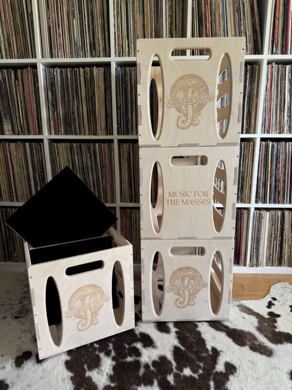 Four Baltic birch 12″ vinyl record crates with Snake Park Records branding—three neatly stacked showing the cobra logo and “Music for the Masses” engraving, and one open crate holding black-sleeved LPs—on a cowhide rug in front of a full white record shelf.