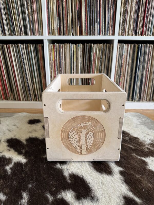 Baltic birch wood 7″ vinyl crate showing laser‑engraved cobra logo on one side, placed on a cowhide rug with vinyl records in the background.