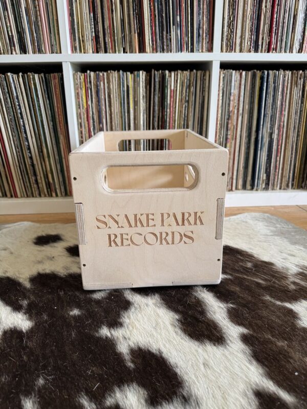 Baltic birch wood 7″ vinyl crate engraved with “Snake Park Records” text on the front, sitting on a cowhide rug in front of a packed white record shelf.