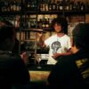 Female bartender shaking a cocktail shaker while wearing a Snake Park Records white T shirt, standing behind a stocked bar with two patrons in the foreground.