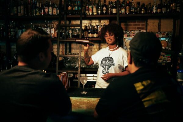 Female bartender shaking a cocktail shaker while wearing a Snake Park Records white T shirt, standing behind a stocked bar with two patrons in the foreground.