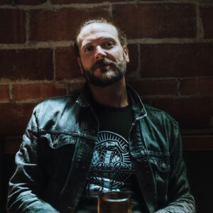 Bearded man in a Snake Park Records black T shirt seated at a table with a green cocktail glass, leaning against a brick wall in a dim venue.