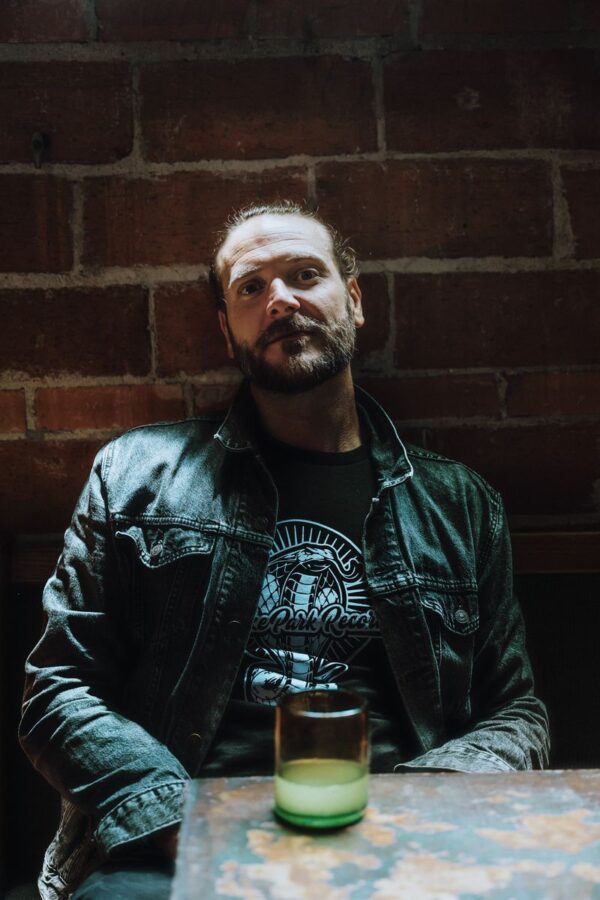Bearded man in a Snake Park Records black T shirt seated at a table with a green cocktail glass, leaning against a brick wall in a dim venue.