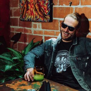 Smiling bearded man wearing a Snake Park Records black T shirt under a denim jacket, holding a green drink while seated against a brick wall.
