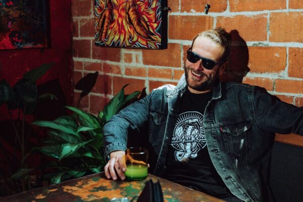 Smiling bearded man wearing a Snake Park Records black T shirt under a denim jacket, holding a green drink while seated against a brick wall.