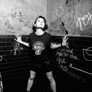 Black and white image of a woman wearing a Snake Park Records black T shirt and leather skirt, standing with her hands on a graffiti‑covered tiled wall.