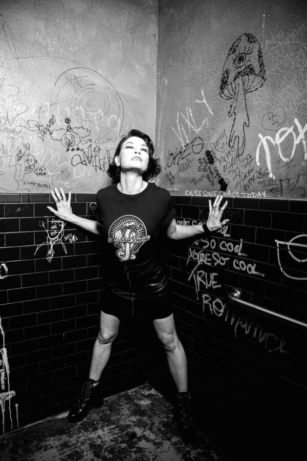Black and white image of a woman wearing a Snake Park Records black T shirt and leather skirt, standing with her hands on a graffiti‑covered tiled wall.
