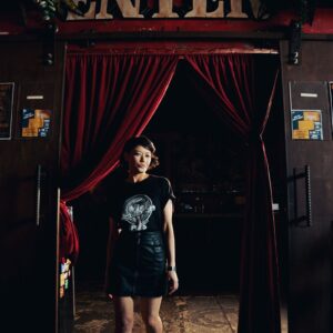 Woman in a Snake Park Records black T shirt and leather skirt walking through a doorway framed by red velvet curtains under an “Enter” sign.