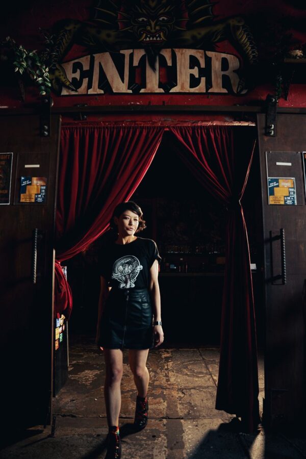 Woman in a Snake Park Records black T shirt and leather skirt walking through a doorway framed by red velvet curtains under an “Enter” sign.