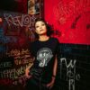 Woman wearing a Snake Park Records black T shirt and black leather skirt, standing against a red and black graffiti‑covered wall in a dimly lit venue.