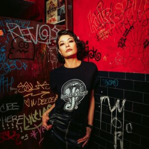 Woman wearing a Snake Park Records black T shirt and black leather skirt, standing against a red and black graffiti‑covered wall in a dimly lit venue.