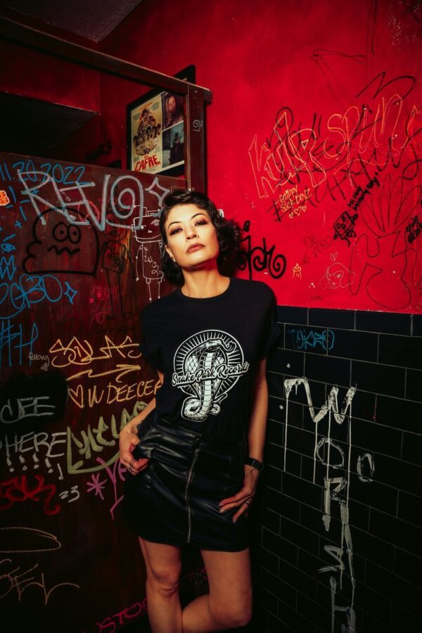 Woman wearing a Snake Park Records black T shirt and black leather skirt, standing against a red and black graffiti‑covered wall in a dimly lit venue.