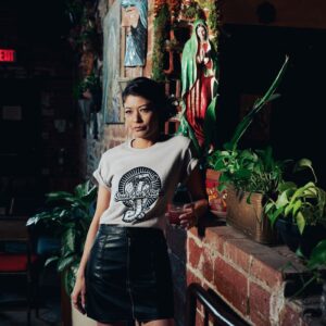 Woman in a Snake Park Records light gray T shirt and black leather skirt holding a cocktail glass, standing against a brick wall adorned with plants and statuettes.
