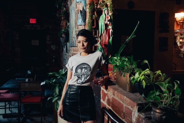 Woman in a Snake Park Records light gray T shirt and black leather skirt holding a cocktail glass, standing against a brick wall adorned with plants and statuettes.