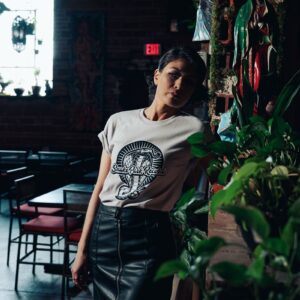 Woman wearing a Snake Park Records light gray T shirt, leaning against a brick ledge covered in potted plants and religious iconography inside a dimly lit bar.