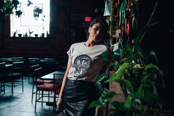 Woman wearing a Snake Park Records light gray T shirt, leaning against a brick ledge covered in potted plants and religious iconography inside a dimly lit bar.