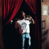 Man wearing a Snake Park Records light gray T shirt and jeans standing in a doorway framed by red velvet curtains, smiling as he holds one curtain aside.