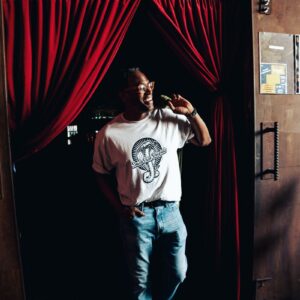 Man wearing a Snake Park Records light gray T shirt and jeans standing in a doorway framed by red velvet curtains, smiling as he holds one curtain aside.
