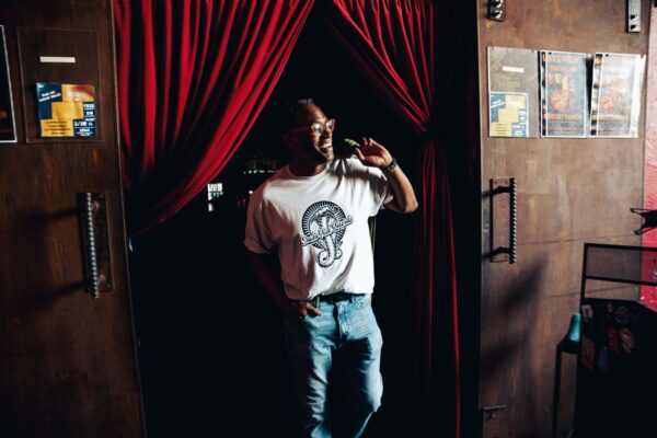 Man wearing a Snake Park Records light gray T shirt and jeans standing in a doorway framed by red velvet curtains, smiling as he holds one curtain aside.