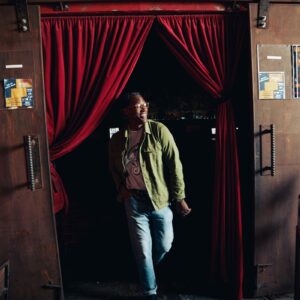 Man in a Snake Park Records light gray T shirt under an olive green overshirt stepping through a doorway with red curtains, jeans rolled at the cuffs.