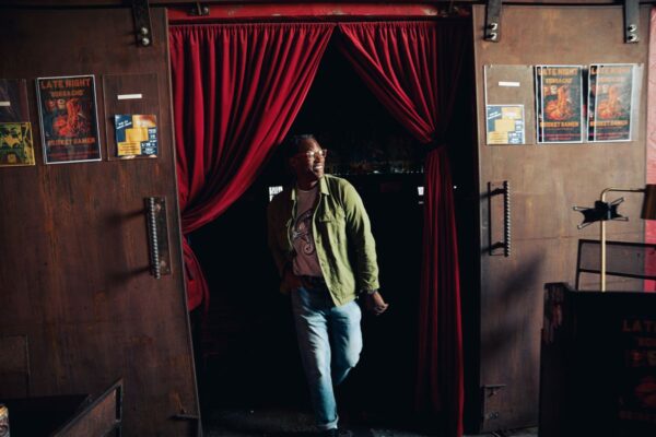 Man in a Snake Park Records light gray T shirt under an olive green overshirt stepping through a doorway with red curtains, jeans rolled at the cuffs.