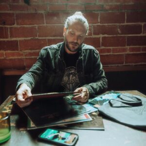 Bearded man in a Snake Park Records black T shirt and denim jacket browsing 12″ vinyl records at a wooden table, with a gray Snake Park Records tote bag and brick wall in the background.