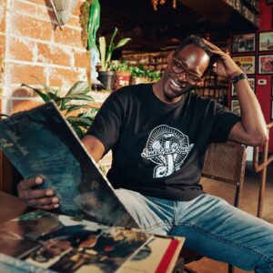 Smiling man wearing a Snake Park Records black T shirt and glasses seated at a table, holding a 12″ vinyl record in a warmly lit, plant‑filled record‑shop setting.