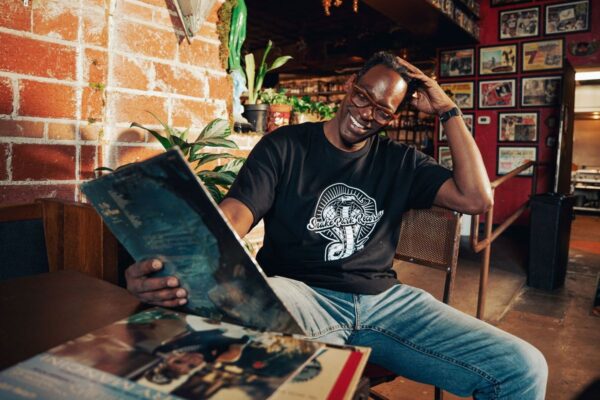 Smiling man wearing a Snake Park Records black T shirt and glasses seated at a table, holding a 12″ vinyl record in a warmly lit, plant‑filled record‑shop setting.