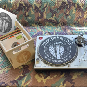 Wooden record crate filled with 12″ and 7″ vinyl next to a Technics turntable fitted with a black Snake Park Records slipmat on a quilted camouflage‑patterned surface.