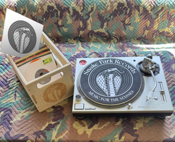 Wooden record crate filled with 12″ and 7″ vinyl next to a Technics turntable fitted with a black Snake Park Records slipmat on a quilted camouflage‑patterned surface.