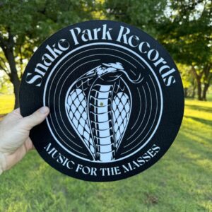 Hand holding a 12″ black slipmat printed with the Snake Park Records cobra logo and “Music for the Masses” text against a sunlit grassy backdrop.
