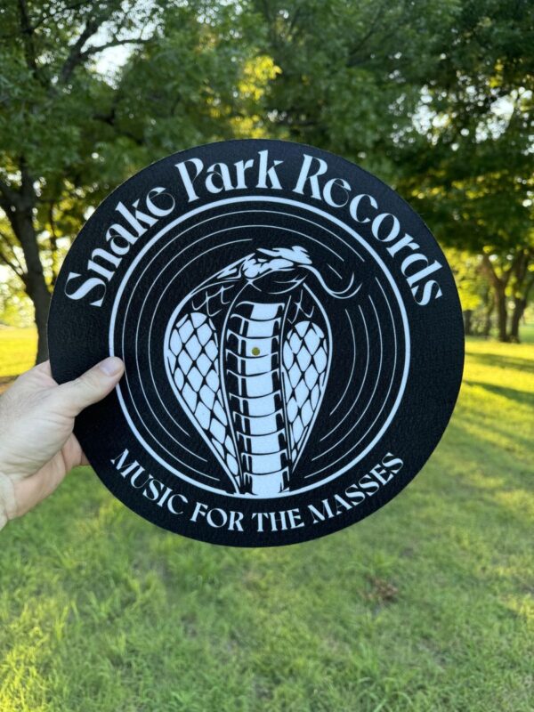 Hand holding a 12″ black slipmat printed with the Snake Park Records cobra logo and “Music for the Masses” text against a sunlit grassy backdrop.