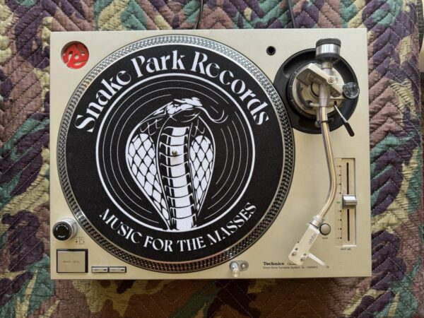 Top‑down view of a Technics SL‑1200 turntable with a Snake Park Records black slipmat featuring a white cobra logo and “Music for the Masses” text.