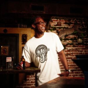 Man wearing a Snake Park Records white T shirt, holding a beer bottle at a dimly lit bar with a brick wall and taxidermy bobcat in the background.