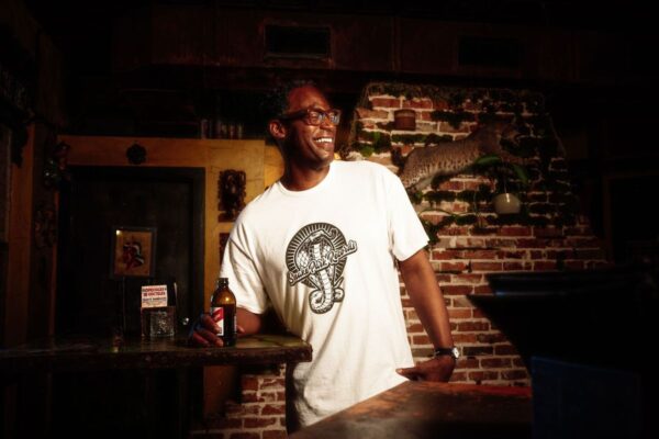 Man wearing a Snake Park Records white T shirt, holding a beer bottle at a dimly lit bar with a brick wall and taxidermy bobcat in the background.