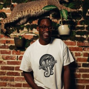 Smiling man in a Snake Park Records white T shirt standing against an exposed brick wall decorated with a mounted bobcat and hanging plants.