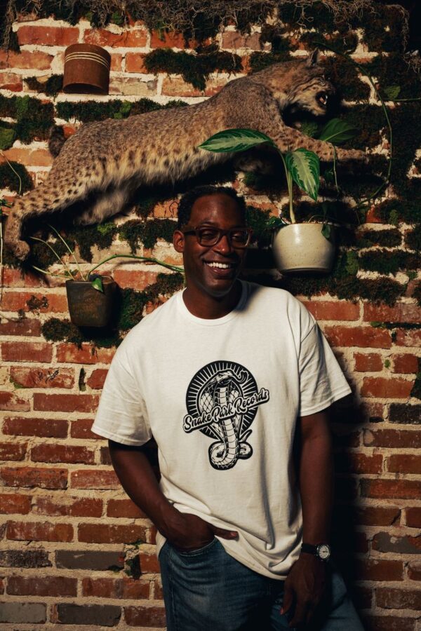 Smiling man in a Snake Park Records white T shirt standing against an exposed brick wall decorated with a mounted bobcat and hanging plants.