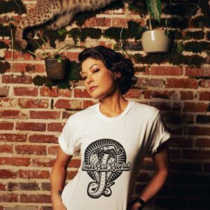 Woman wearing a Snake Park Records white T shirt, tugging at the neckline while standing against a brick wall with a mounted bobcat and hanging plants.