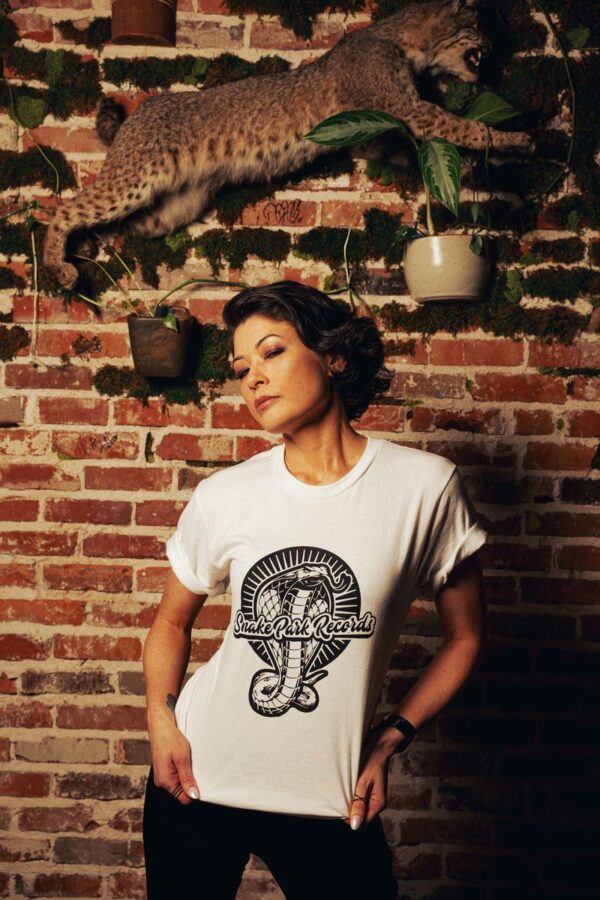 Woman wearing a Snake Park Records white T shirt, tugging at the neckline while standing against a brick wall with a mounted bobcat and hanging plants.