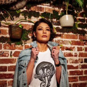 Woman in a Snake Park Records white T shirt slipping on a denim jacket in front of an exposed brick wall decorated with greenery and a taxidermy bobcat.