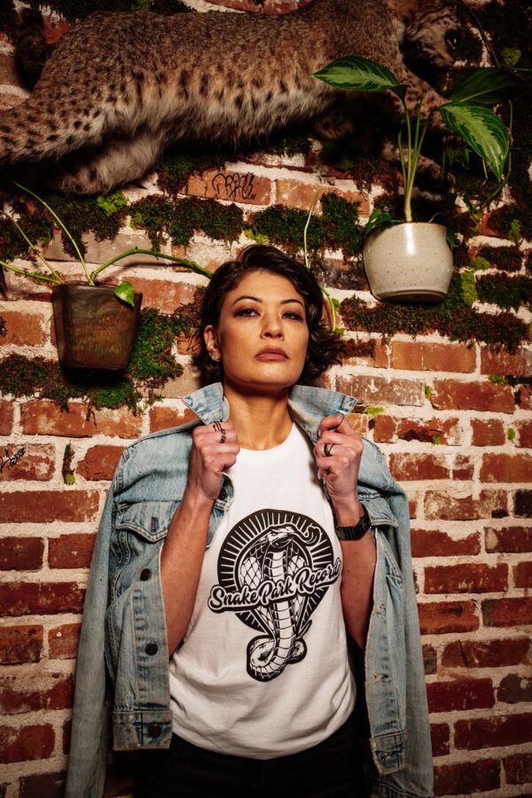 Woman in a Snake Park Records white T shirt slipping on a denim jacket in front of an exposed brick wall decorated with greenery and a taxidermy bobcat.
